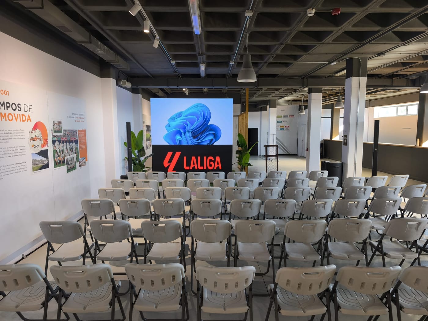 Pantalla LED encendida al fondo de una sala interior con las sillas preparadas para el público