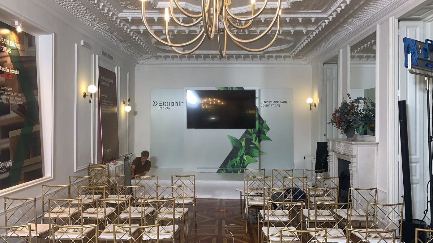 Televisor sobre soporte al fondo de un salón con sillas preparadas para una presentación de empresa