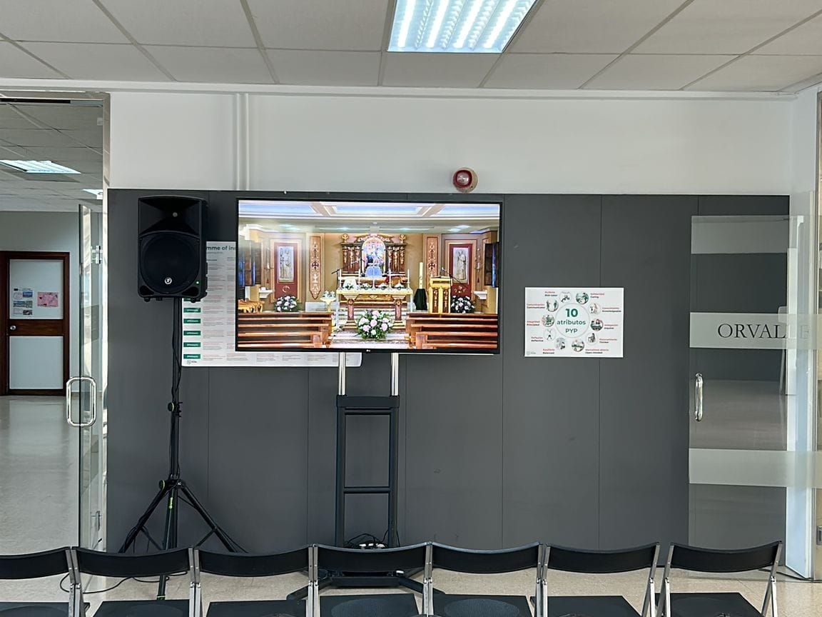 Televisor de gran formato sobre soporte de suelo, presidiendo una sala con las sillas colocadas para una formación