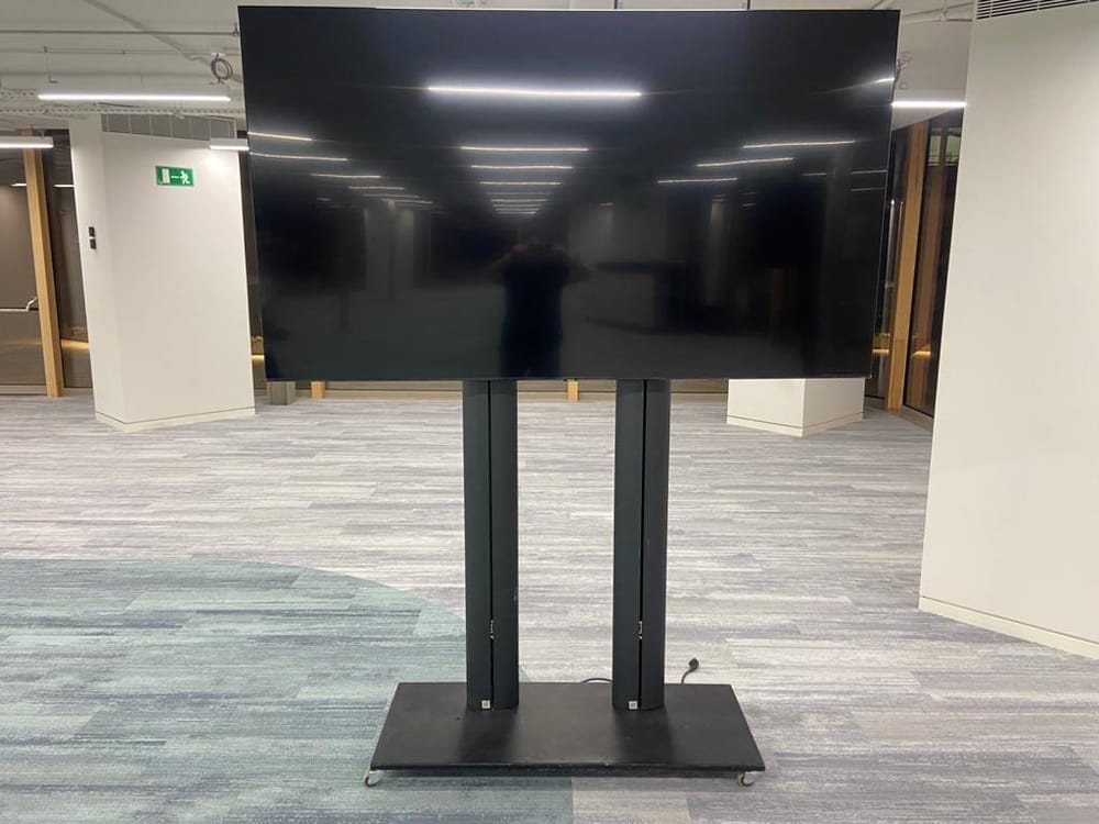 Televisor montado sobre un soporte de suelo de doble columna con base plana, en el pasillo de un espacio de oficinas